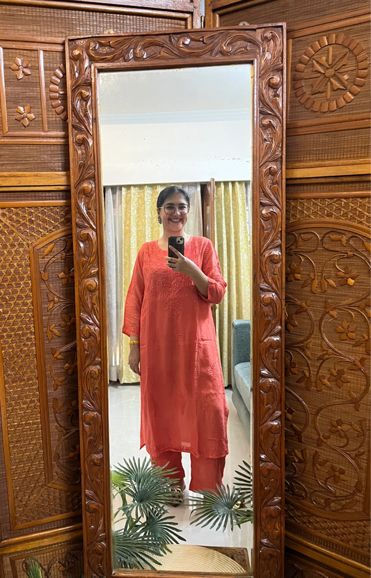 Person wearing a coral kurta standing in front of a decorative wooden mirror.